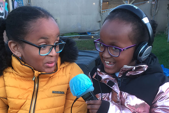 2 young sisters sat on bench, one interviewing the other.