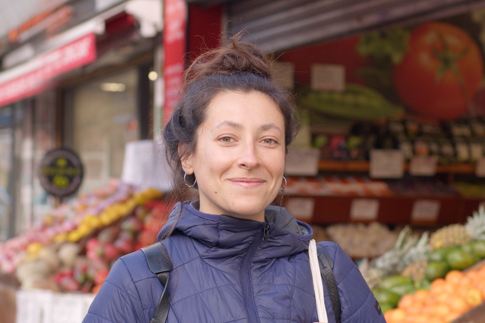 Photo shows woman wearing a rucksack, stood in front of a fruit and veg shop, the fruit displayed colourfully in the background. Awnings and shutters also visible in background looks like a row of shops