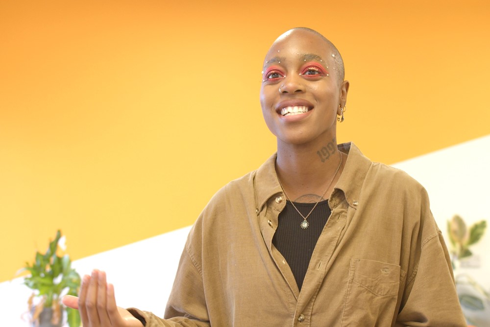 Photo shows woman, gesturing as if speaking, and smiling, with office wall and office plants as background