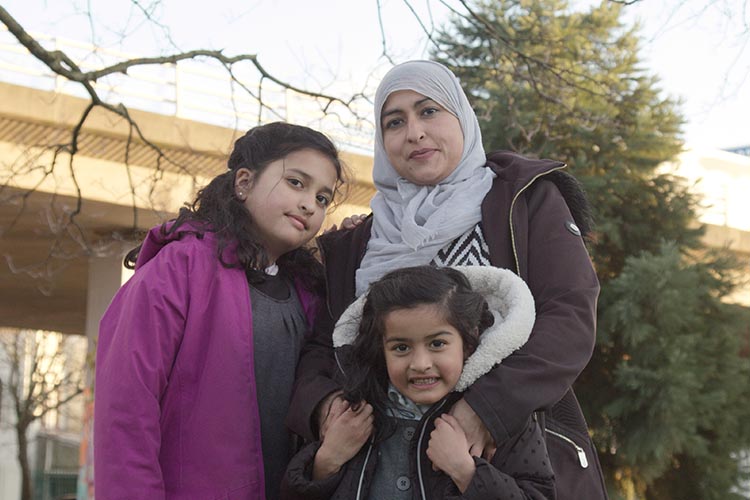 Photo shows family stood together – woman with headscarf has her arms around her younger daughter and the elder daughter is stood next to them. There are trees and a motorway flyover behind them