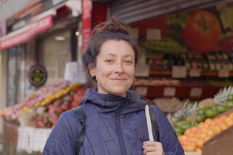 Photo shows woman wearing a rucksack, stood in front of a fruit and veg shop, the fruit displayed colourfully in the background. Awnings and shutters also visible in background looks like a row of shops