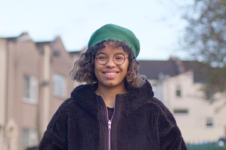 Photo shows young woman smiling with terrace housing in background and a tree and lamppost