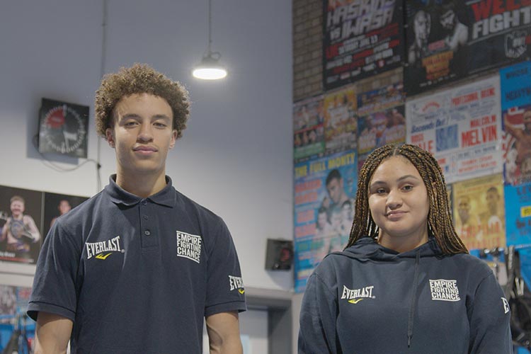 Photo shows a male and female teenager, both wearing Empire Fighting Chance blue tops, in front of gym walls covered in posters for boxing matches / events