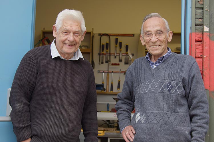 Photo shows two older men stood in doorway of a workshop with woodworking tools visible behind them, mounted on the walls