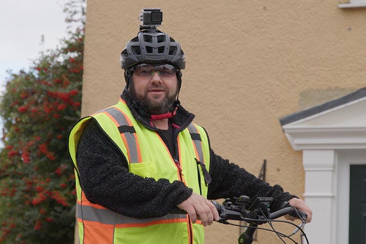 Photo shows man wearing helmet with sports video camera mounted on top, stood in front of a house with his bike