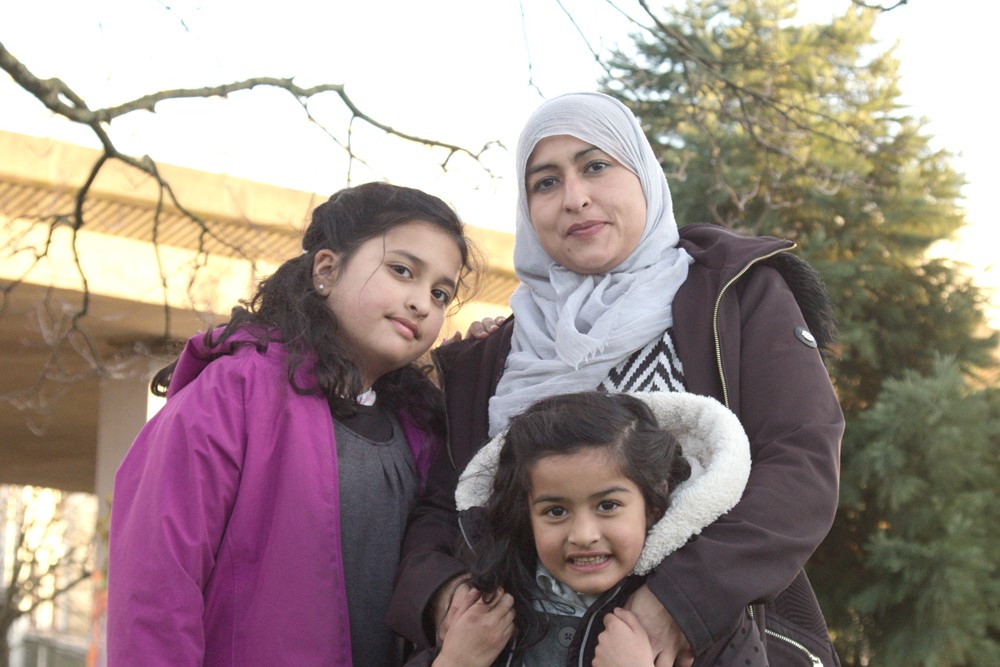 Photo shows family stood together – woman with headscarf has her arms around her younger daughter and the elder daughter is stood next to them. There are trees and a motorway flyover behind them