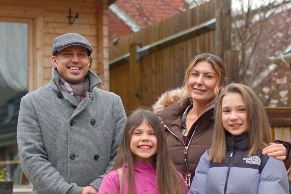 Photo shows smiling family group, man, woman and two daughters stood in the garden of a house, with neighbouring houses in background