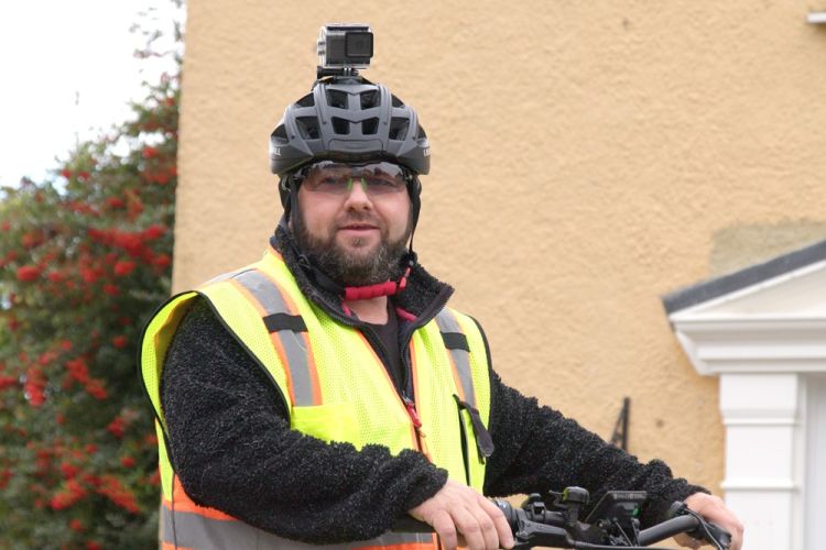 Photo shows man wearing helmet with sports video camera mounted on top, stood in front of a house with his bike