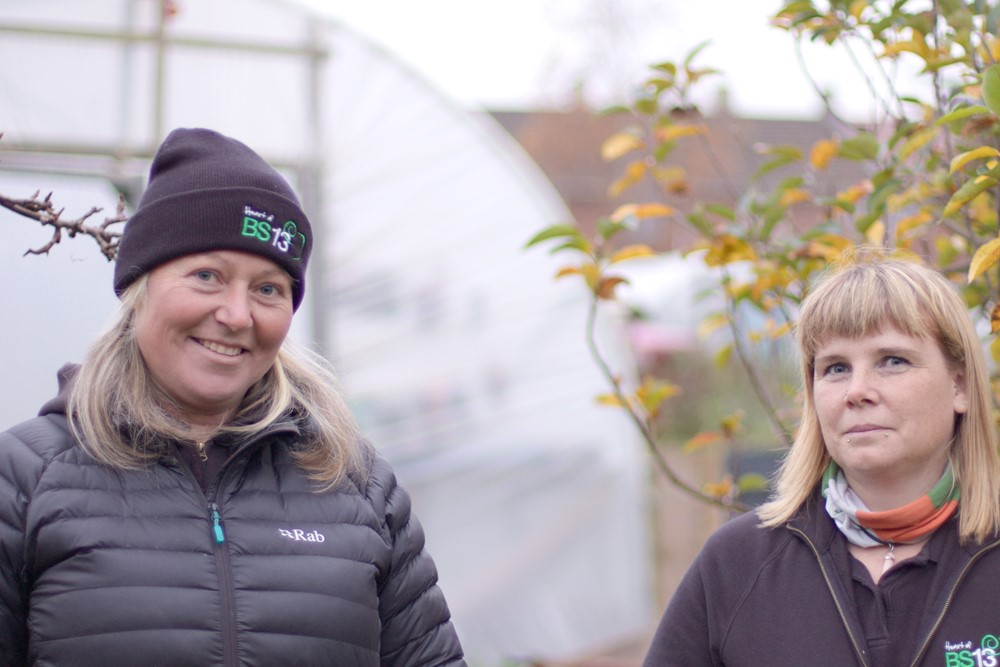 Photo shows two women in Heart of BS13 branded clothing with polytunnels and fruit trees in the background