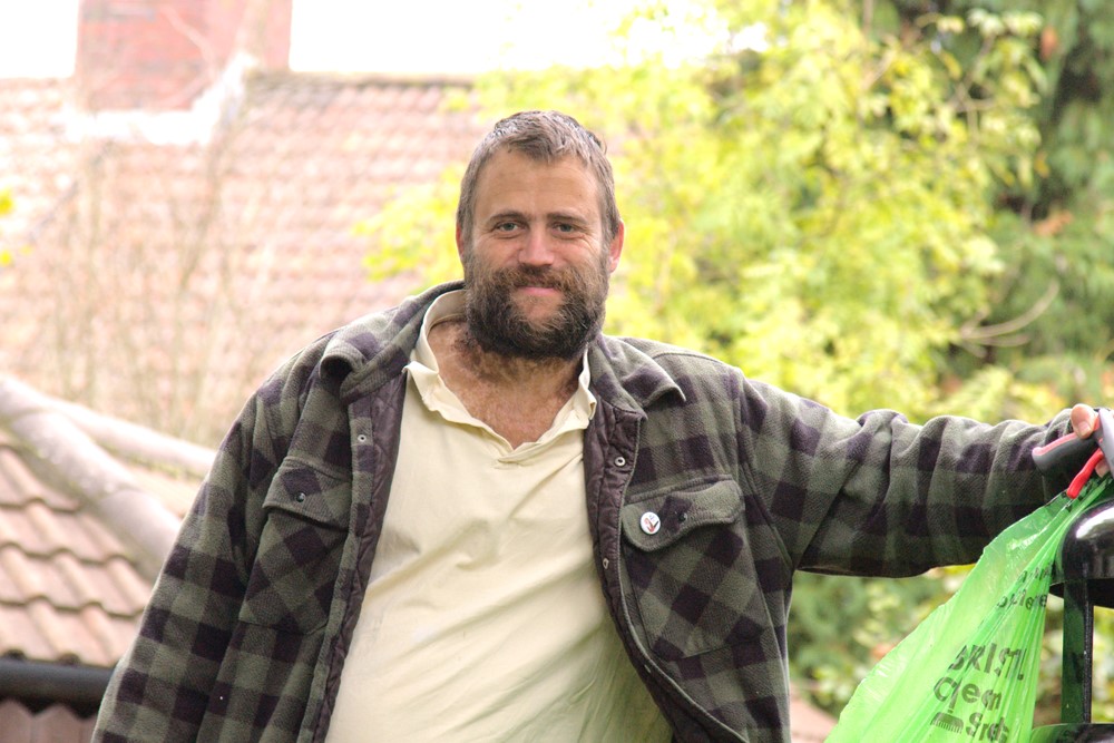 Photo shows man leaning on a bin, holding a litter-picking stick and Bristol Waste sac in left hand, with trees and houses in background