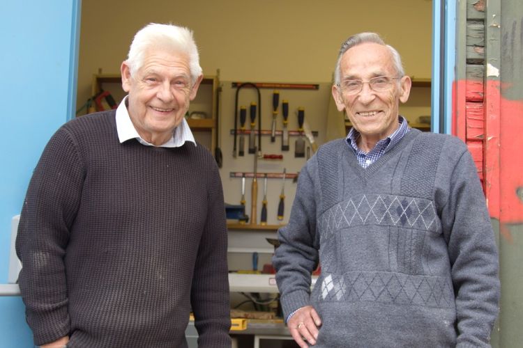 Two men standing next to each other smiling, in front of a tool shed.