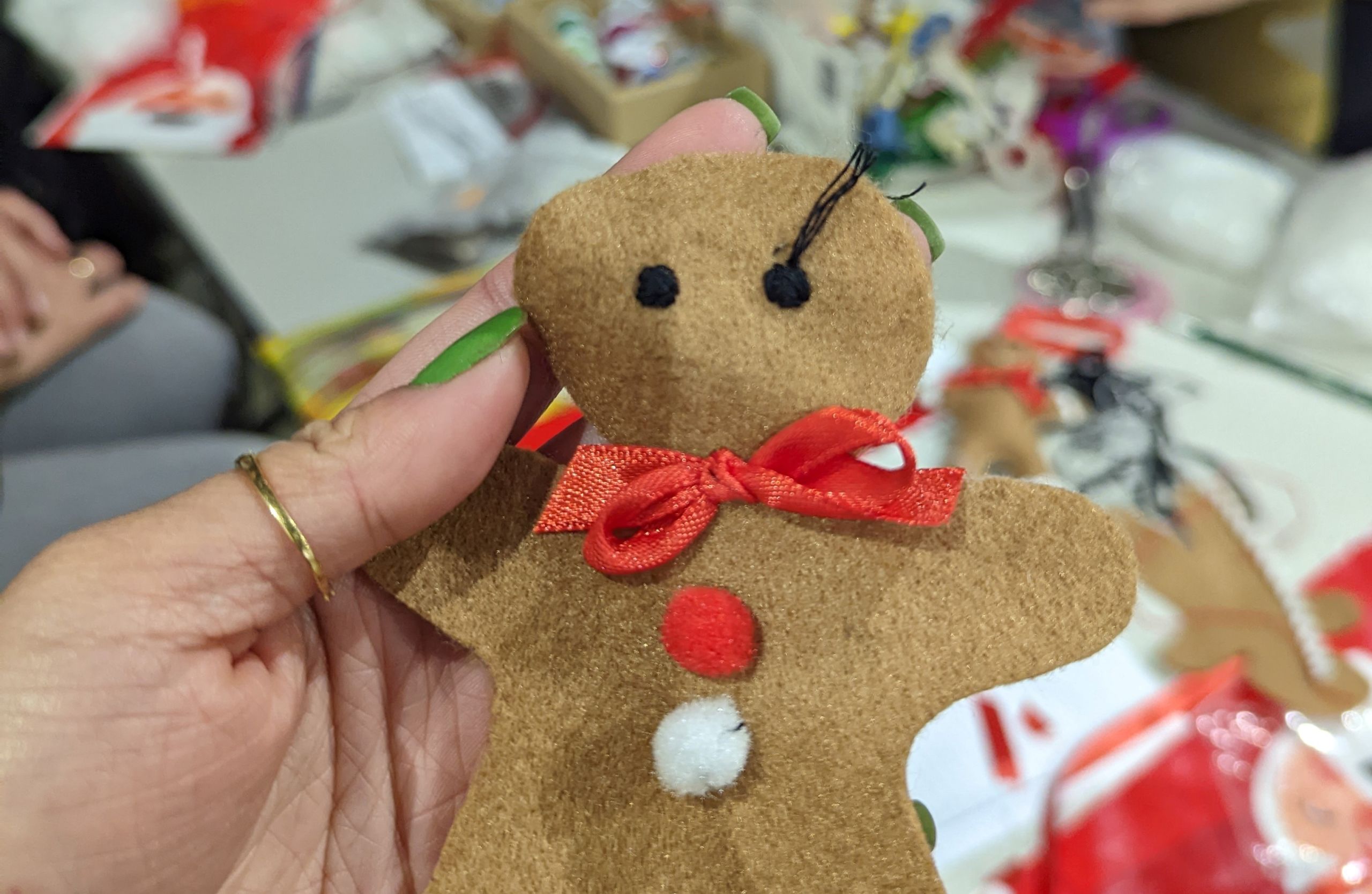 Gingerbread tree decoration craft workshop person sewing a fabric 'gingerbread' person tree decoration at craft workshop. The decoration has a red bow and two 'buttons' one red, one white. In the background are the tools and materials for making fabric decorations.